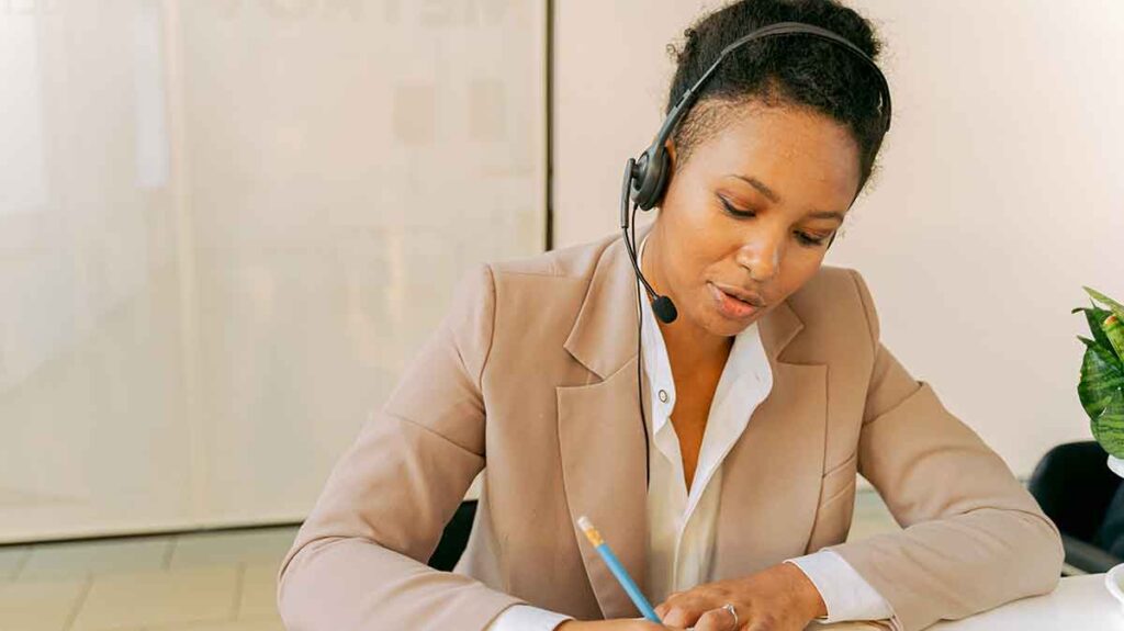 Female telephone interpreter sitting at a desk. She's looking down and taking notes while interpreting over the phone.