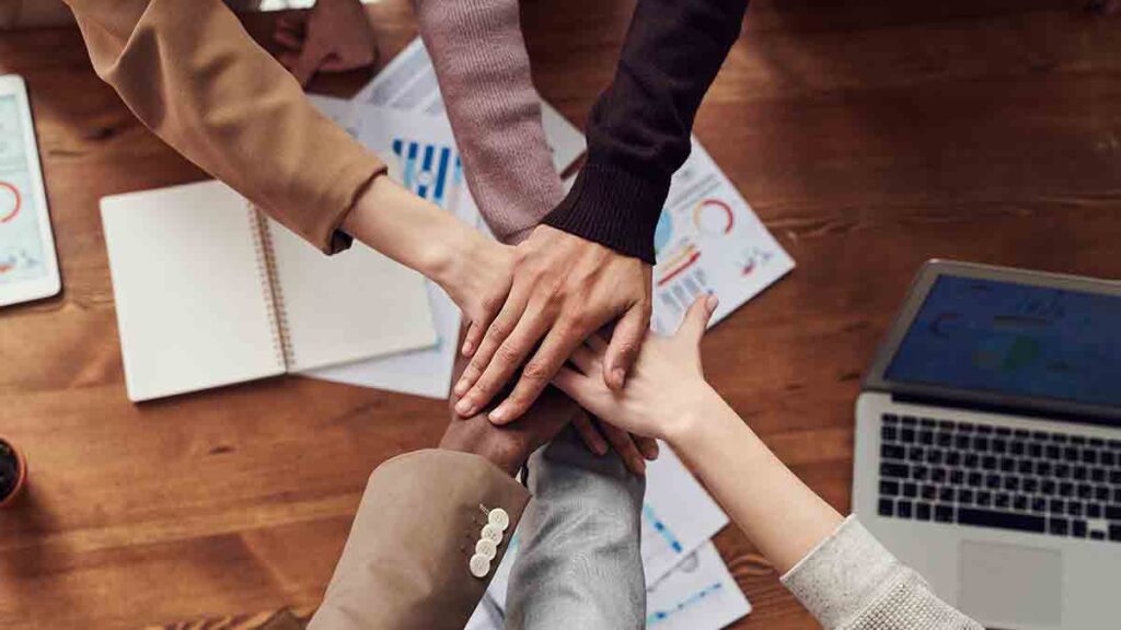 Six interpreting industry professionals put their hands together in a hand-stack over a table that has papers and a laptop on it. Their faces are not shown.