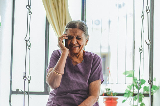 A smiling elder talks on the phone while sitting outside on a balcony.