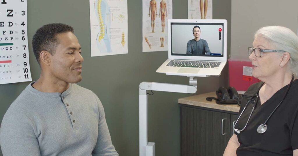 A nurse talks to a patient with the help of a video interpreter who speaks through a laptop stationed on a rolling stand.